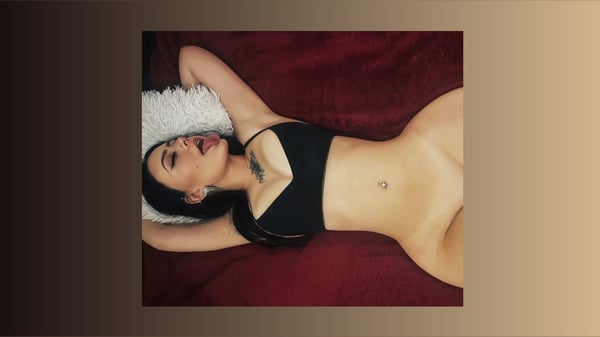 A young woman lying on a red surface with pillows, wearing a black top, posing confidently with tattoos and a navel piercing visible.