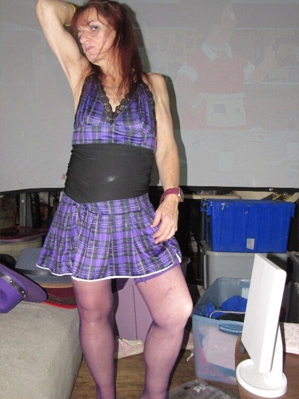A woman with long red hair stands wearing a purple plaid mini dress and sheer stockings, posing confidently in front of a white projector screen.