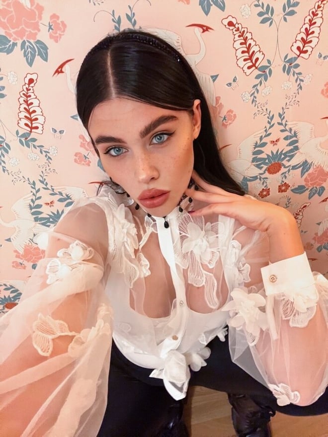 A woman with long dark hair posing in front of a floral-patterned wall, wearing a sheer lace blouse and choker necklace.