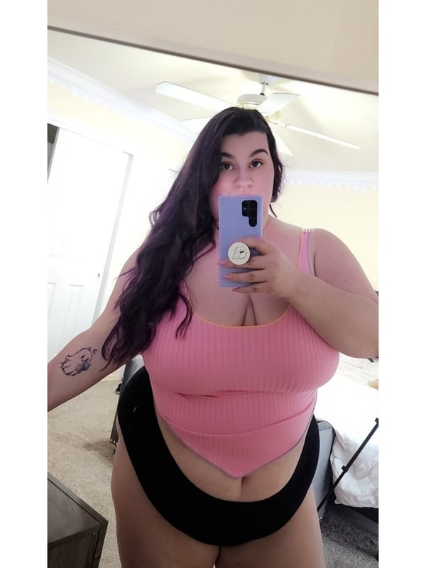 A woman with long dark hair posing in front of a mirror, wearing a pink tank top and black bottoms, holding her phone.