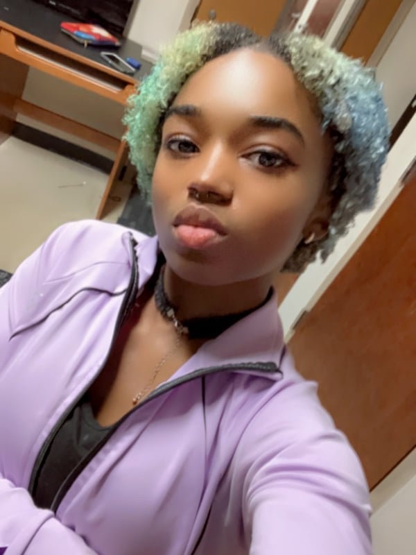 A young woman with colorful dyed hair posing confidently indoors, wearing a purple zip-up jacket and choker necklace.
