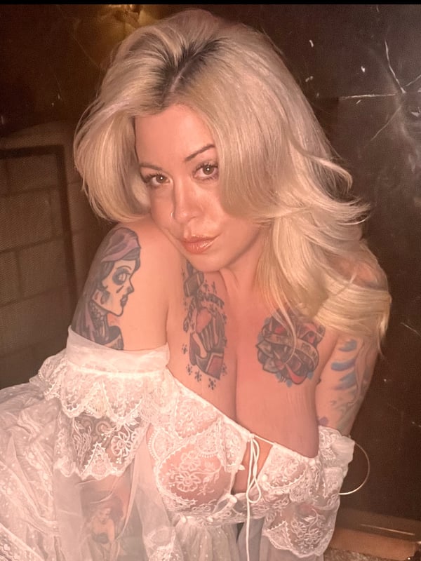 A woman with long blonde hair and visible tattoos wearing a lace top, posing in a dimly lit room with warm lighting.