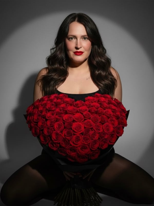 A woman with long wavy dark hair sitting and holding a large bouquet of red roses against her chest, dressed in black attire in a studio-lit background.