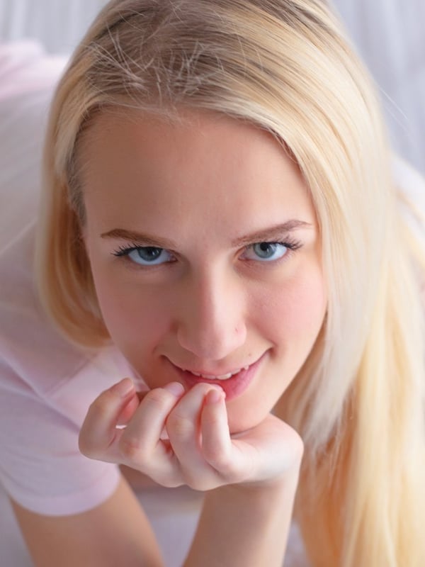 Close-up of a smiling young woman with long blonde hair and blue eyes, wearing a white top, lying on a bed with tattoos on her arm.