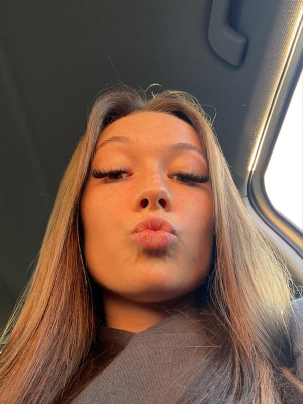 Close-up selfie of a young woman with long blonde hair making a playful pout in a car interior with a window and door visible.
