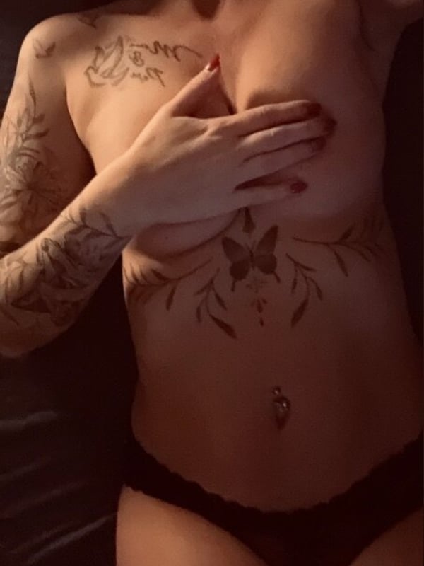 A woman with tattoos poses topless, covering her chest with her hands while wearing black underwear, in a dimly lit indoor setting.