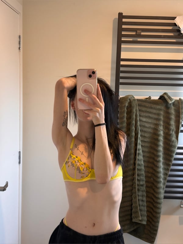 A woman taking a mirror selfie in a yellow top and dark bottoms in a bathroom setting.