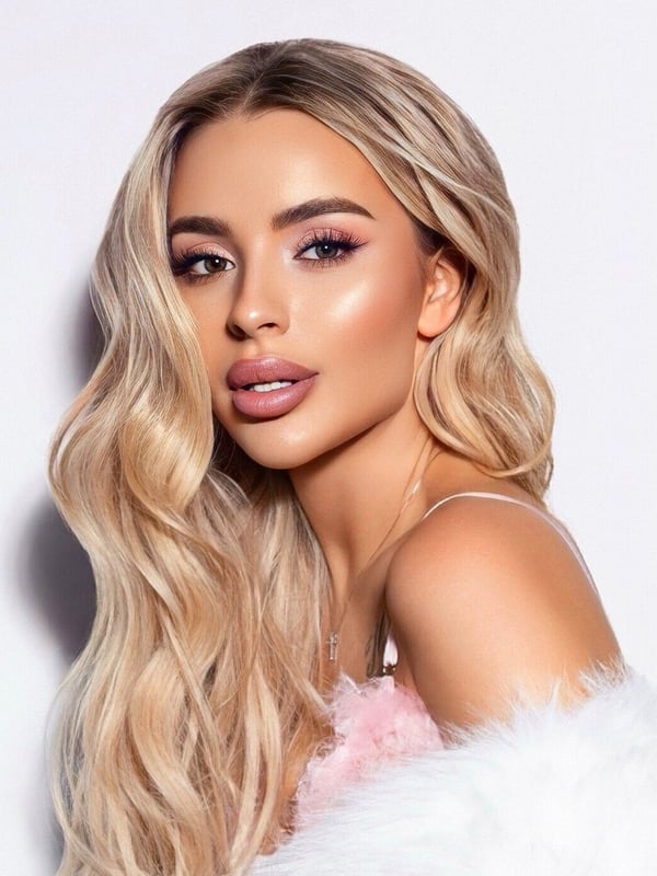 A woman with long blonde hair, glamorous makeup, and a fur-trimmed top, posing confidently against a light background.