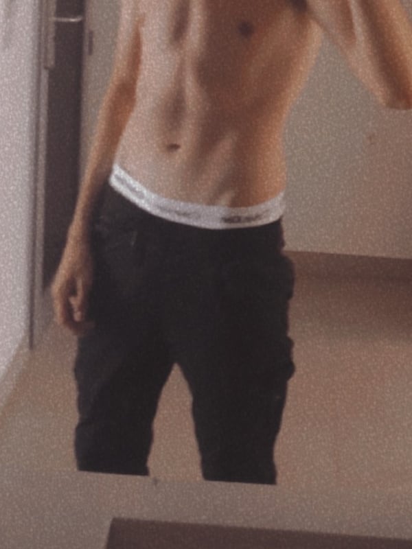 Young man posing shirtless in underwear for a mirror selfie in an indoor setting.