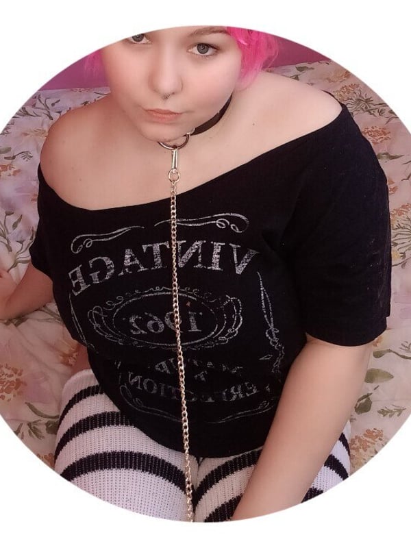 A woman with pink hair wearing an off-shoulder black top and choker necklace, sitting on a bed with a floral background.