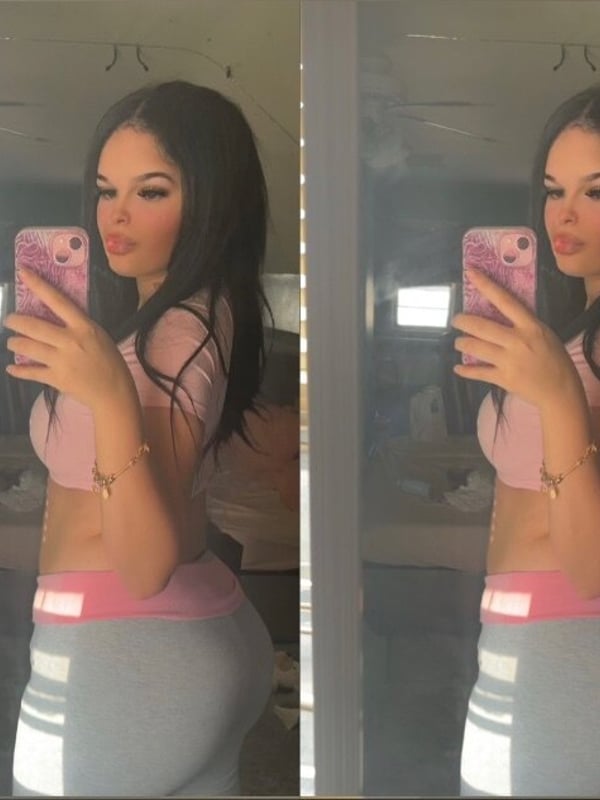 Young woman taking a mirror selfie in a pink top and gray leggings, showing a confident smile.