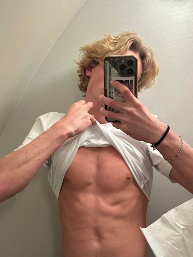 Young man with curly blonde hair taking a mirror selfie in a locker room, wearing a white t-shirt and grey sweatpants.