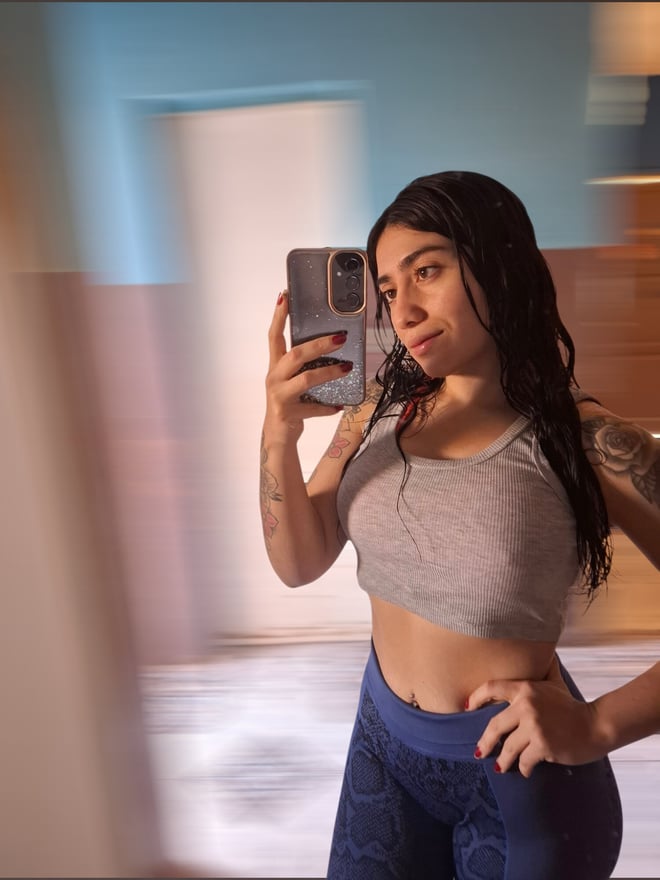 A woman with long dark hair taking a mirror selfie in a gray crop top and blue leggings, standing in a room with colorful lighting.