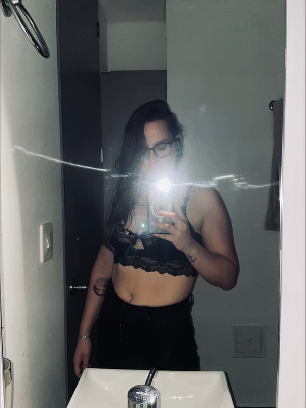 A woman with glasses and long hair stands in front of a bathroom mirror, wearing lace lingerie and smiling at the camera.