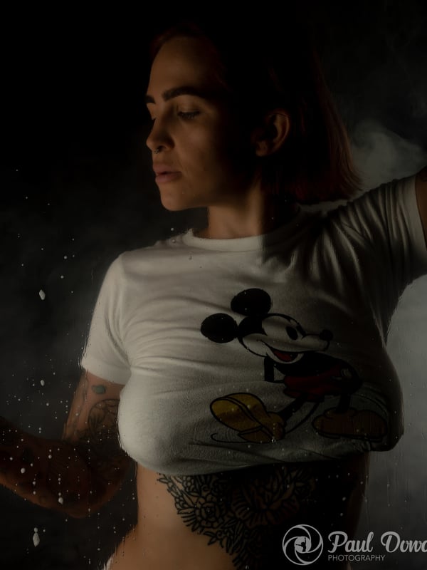 A woman with red hair posing sideways in a white Mickey Mouse t-shirt against a misty background.