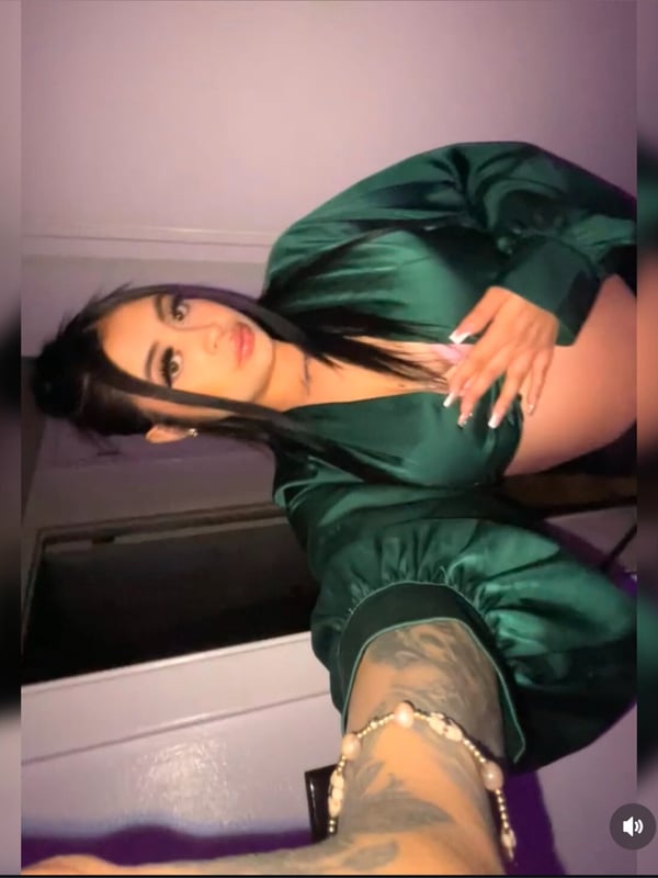 Young woman with long dark hair posing in a shiny green satin outfit, smiling confidently indoors with tattoos visible on her arm.