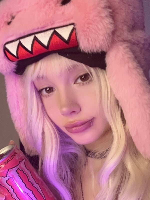 A person wearing a pink furry monster hat and holding a pink energy drink, illuminated with purple lighting.