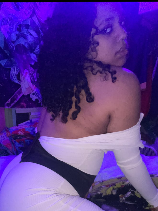 A young woman with curly hair and a lip piercing posing confidently in a dimly lit bedroom with vibrant wall art.