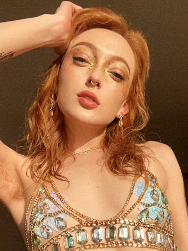 A young woman with red wavy hair posing confidently, wearing a sparkling green and blue beaded top, with tattoos visible on her arm.