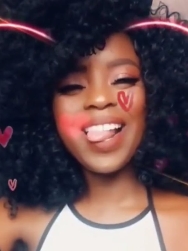 Young Black woman with voluminous curly black hair, smiling playfully with tongue out, adorned with pink heart filters and wearing a white top.