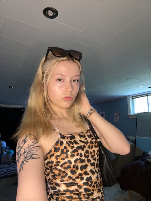 Young blonde woman with sunglasses on her head wearing a leopard print top, standing indoors with a neutral expression and visible arm tattoo.