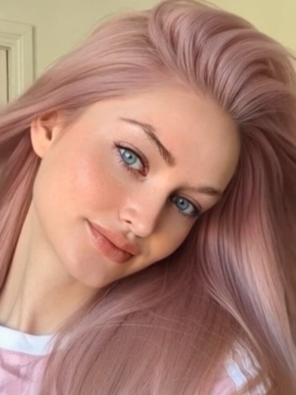 Close-up portrait of a person with long pink hair, blue eyes, and subtle makeup, smiling softly.