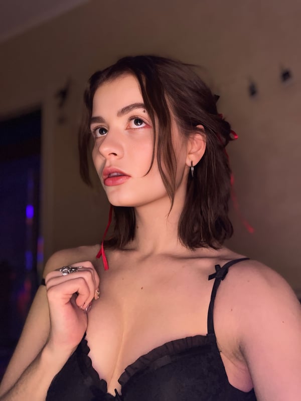 A young woman with brown hair styled in a bun and red ribbons poses confidently in black lace lingerie against a dimly lit indoor setting.