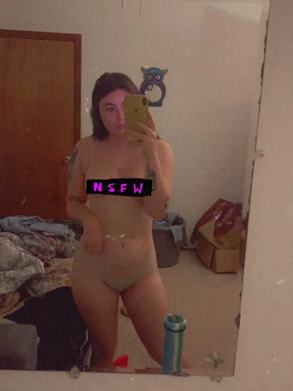 A young woman taking a mirror selfie in a bedroom, wearing minimal clothing with 'NSFW' text across her chest.
