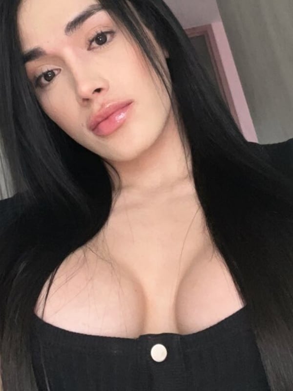 Close-up selfie of a young woman with long dark hair, smiling softly while wearing a black top, indoors with a pink door in the background.