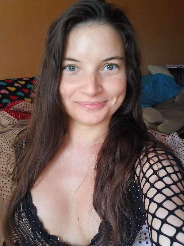 A woman with long brown hair smiling at the camera, wearing a black mesh top, posing in a cozy bedroom with colorful bedding.