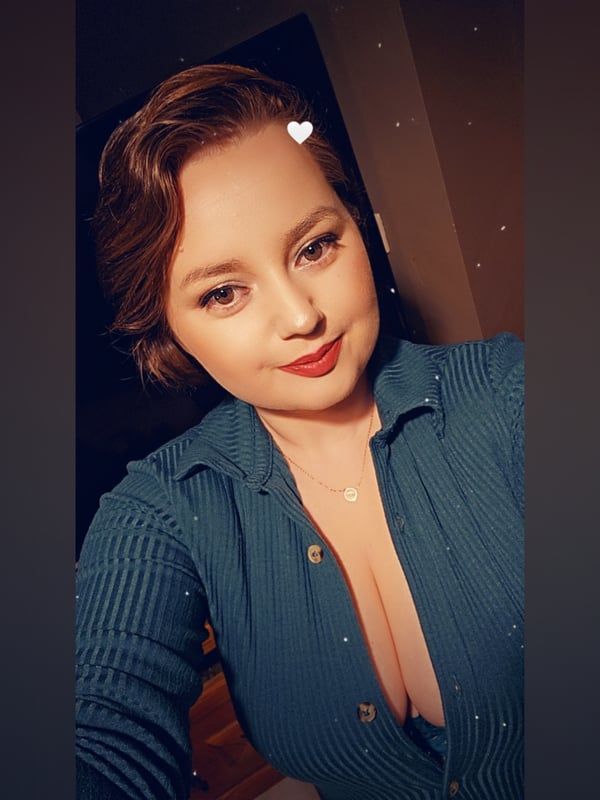 Close-up selfie of a smiling woman with wavy brunette hair, wearing a teal collared top with a deep neckline, against a dark indoor background.