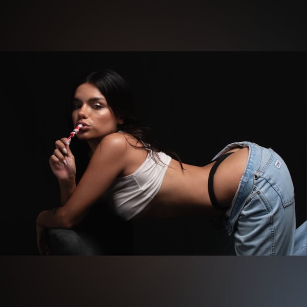 Young woman with long dark hair posing playfully on the floor in a white top and low-rise jeans, holding a red and white striped candy.