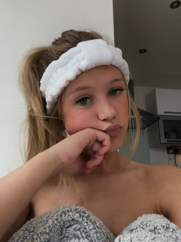 A young woman with blonde hair in a ponytail and white headband, pouting playfully while wrapped in a fuzzy blanket indoors.