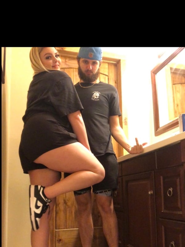 Young woman with blonde hair and man posing playfully together in a bathroom, wearing casual black athletic clothing and sneakers.