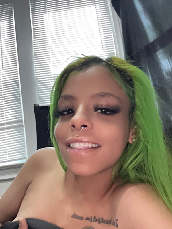 Young woman with green hair smiling at the camera indoors near a window, showing facial piercings and a tattoo on her chest.