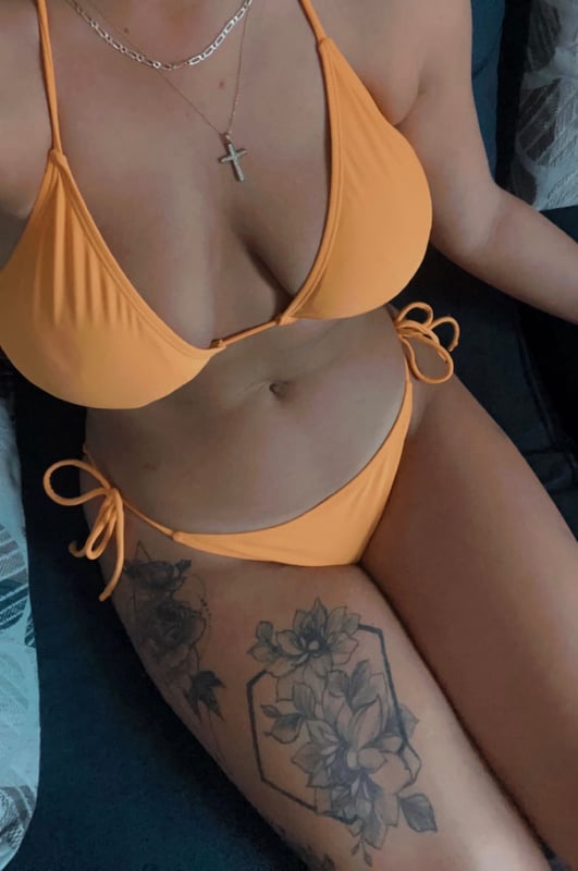 Woman posing in an orange bikini on a couch, showcasing tattoos on her thigh.