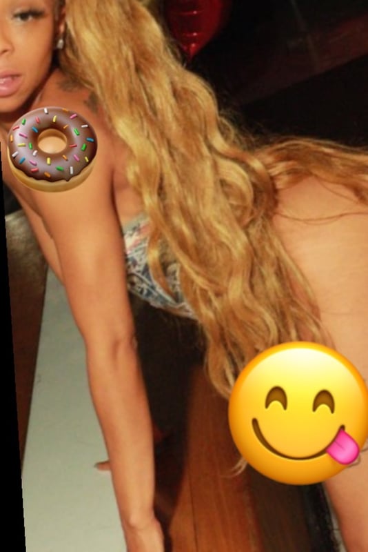 A woman with long blonde hair posing playfully with donuts on her body, smiling with emojis in a vibrant indoor setting.