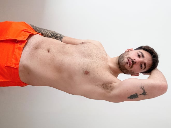 A man lying on his side wearing orange work pants, posing with tattoos visible on his arms against a plain background.