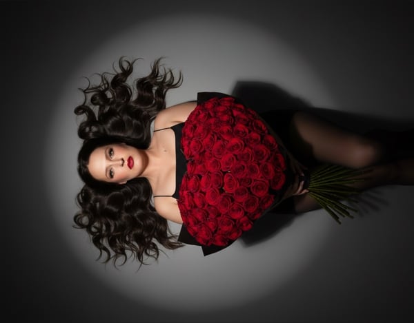 A woman with long wavy dark hair lying on the ground holding a large bouquet of red roses, wearing a black top and stockings in a dimly lit setting.