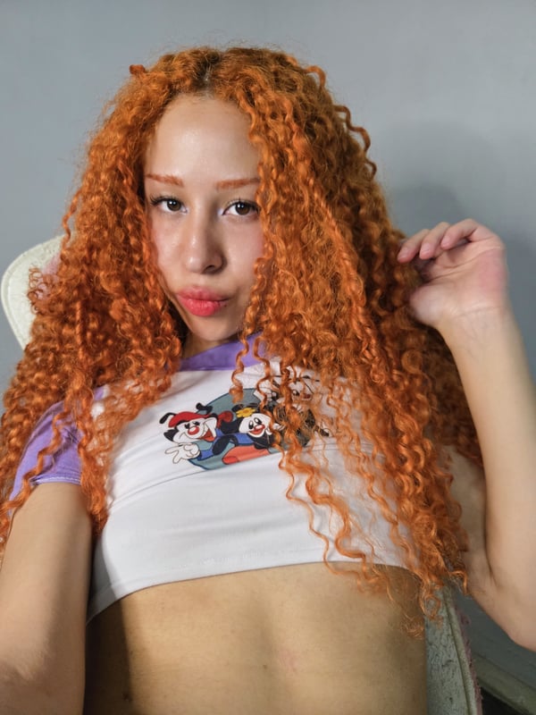 A person with long curly red hair wearing a dragon-print top and jeans, posing indoors.
