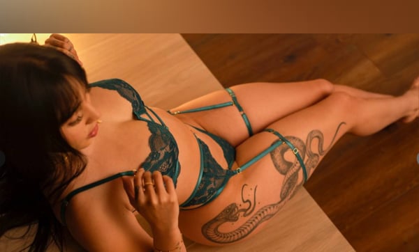 A woman with long dark hair reclining on a wooden floor, wearing green lingerie and displaying intricate tattoos on her body.