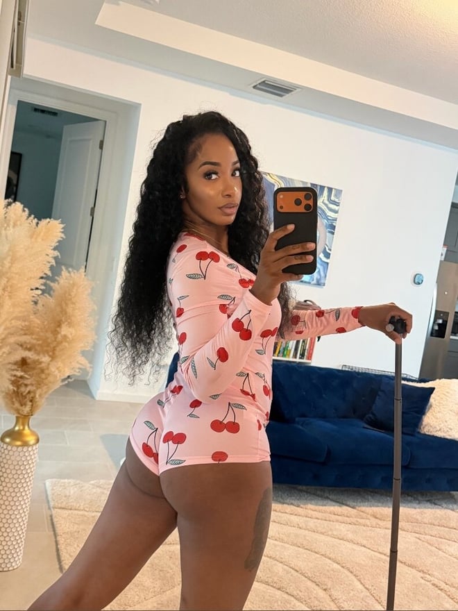 Young woman with long wavy dark hair taking a mirror selfie in a pink cherry-patterned pajama set, standing in a stylish bedroom with plants and furniture.