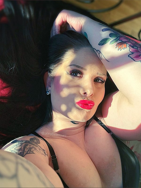 A woman with dark hair, tattoos, and piercings lies on a surface in sunlight, wearing a black top and making a playful expression.