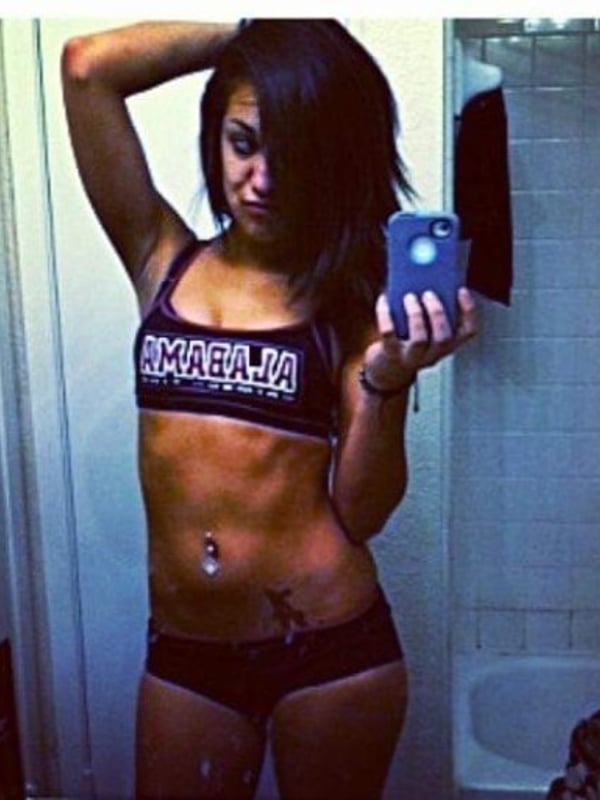 A fit young woman posing confidently in a sports bra and shorts in front of a mirror, holding a phone.