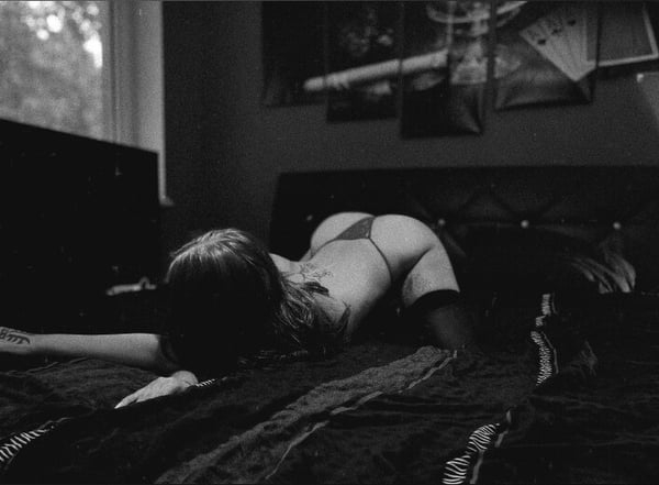 A woman lying on a bed in a dimly lit room with dark decor and artwork on the walls, wearing lingerie and stockings.