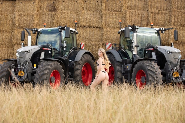 A blonde woman posing playfully between two large tractors in a hay field with bales in the background.