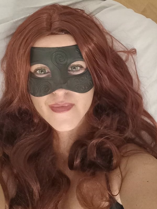 A woman lying on a bed wearing a black mask and red wavy hair, smiling at the camera.