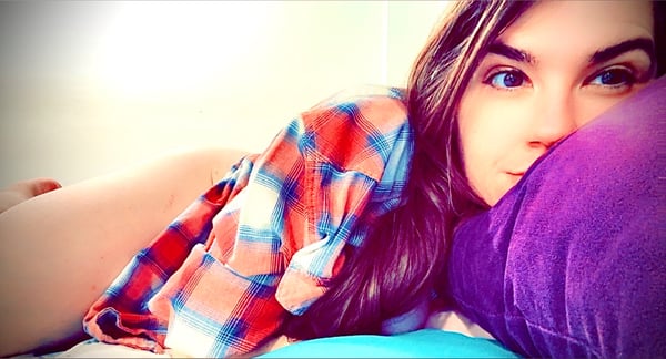 A woman with long brown hair leans on colorful pillows, wearing a plaid shirt, smiling playfully in a cozy indoor setting.