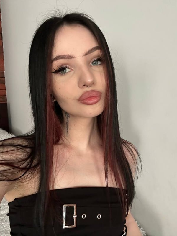 Young woman with long dark hair with red highlights, posing confidently in a black top against a bedroom background.