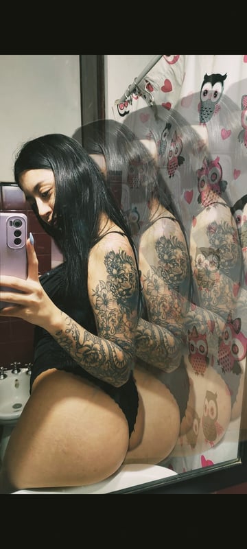 A woman with long dark hair and tattoos poses in a black outfit in front of a mirror with decorative wall art.